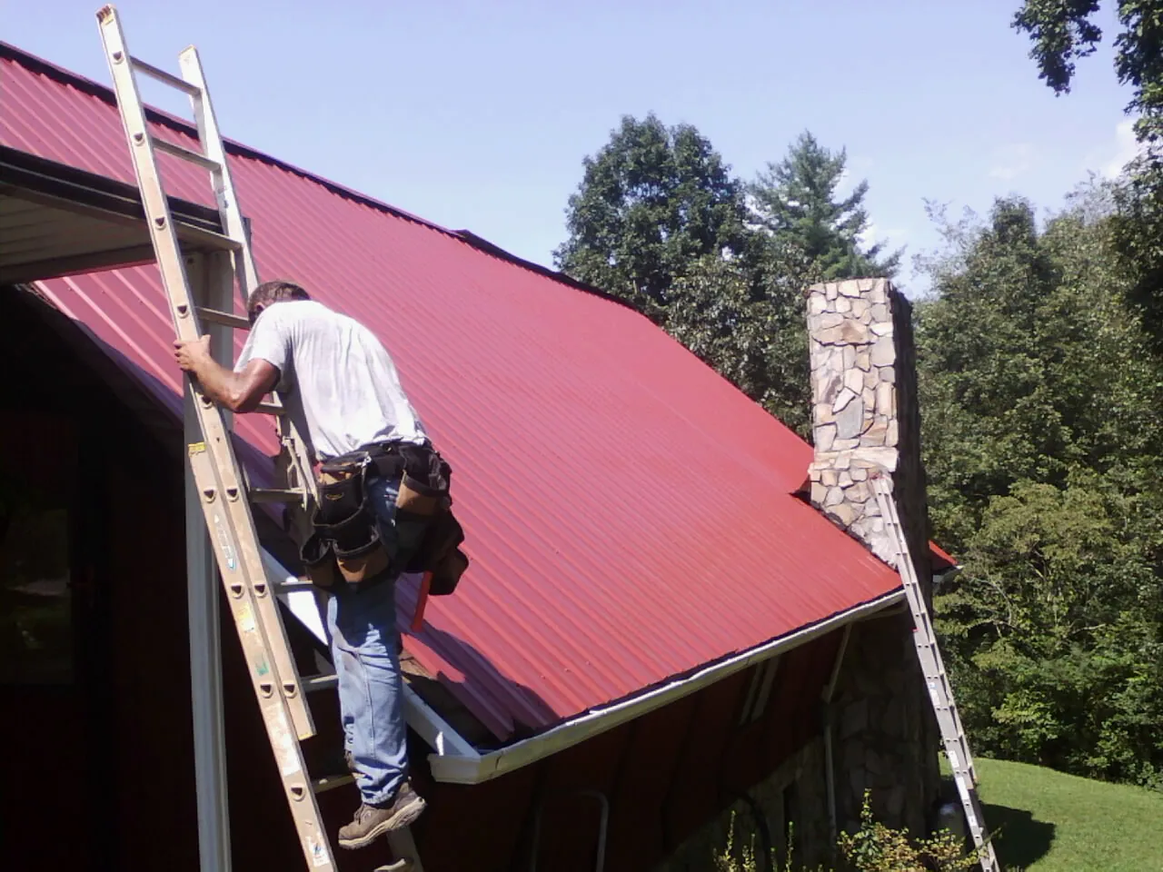 Roofer on a ladder beside a red metal roof and stone chimney — Soffit & Fascia Repair by Roofing Brothers in Moroni